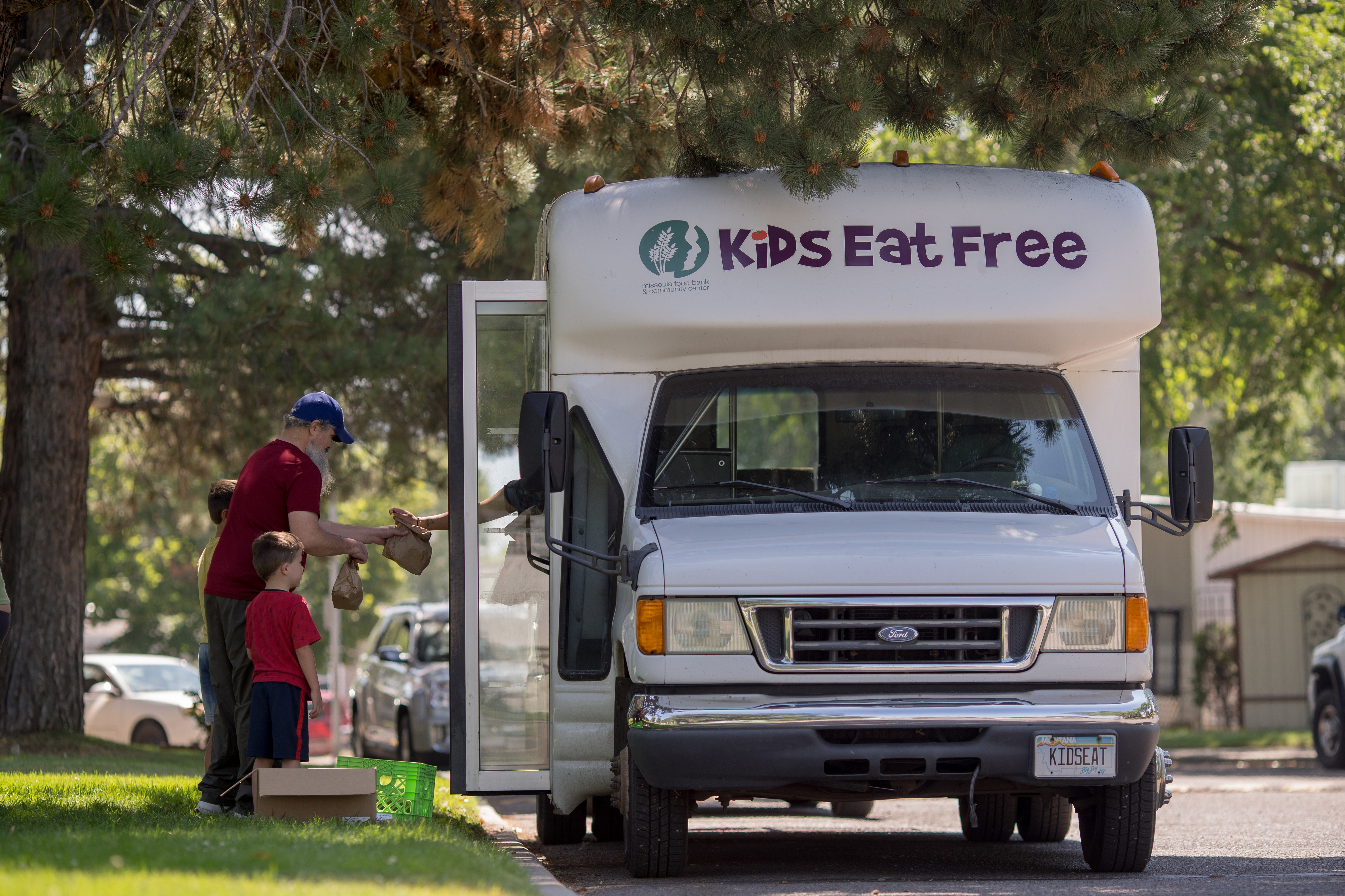 Kids line up at a park to get meals from a small white bus that says "Kids Eat Free" on the top. 
