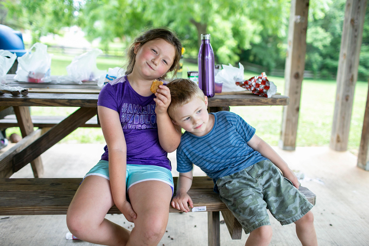 A brother and sister sit at a picnic table. The brother rests his head on the sisters shoulder while she smiles at the camera.