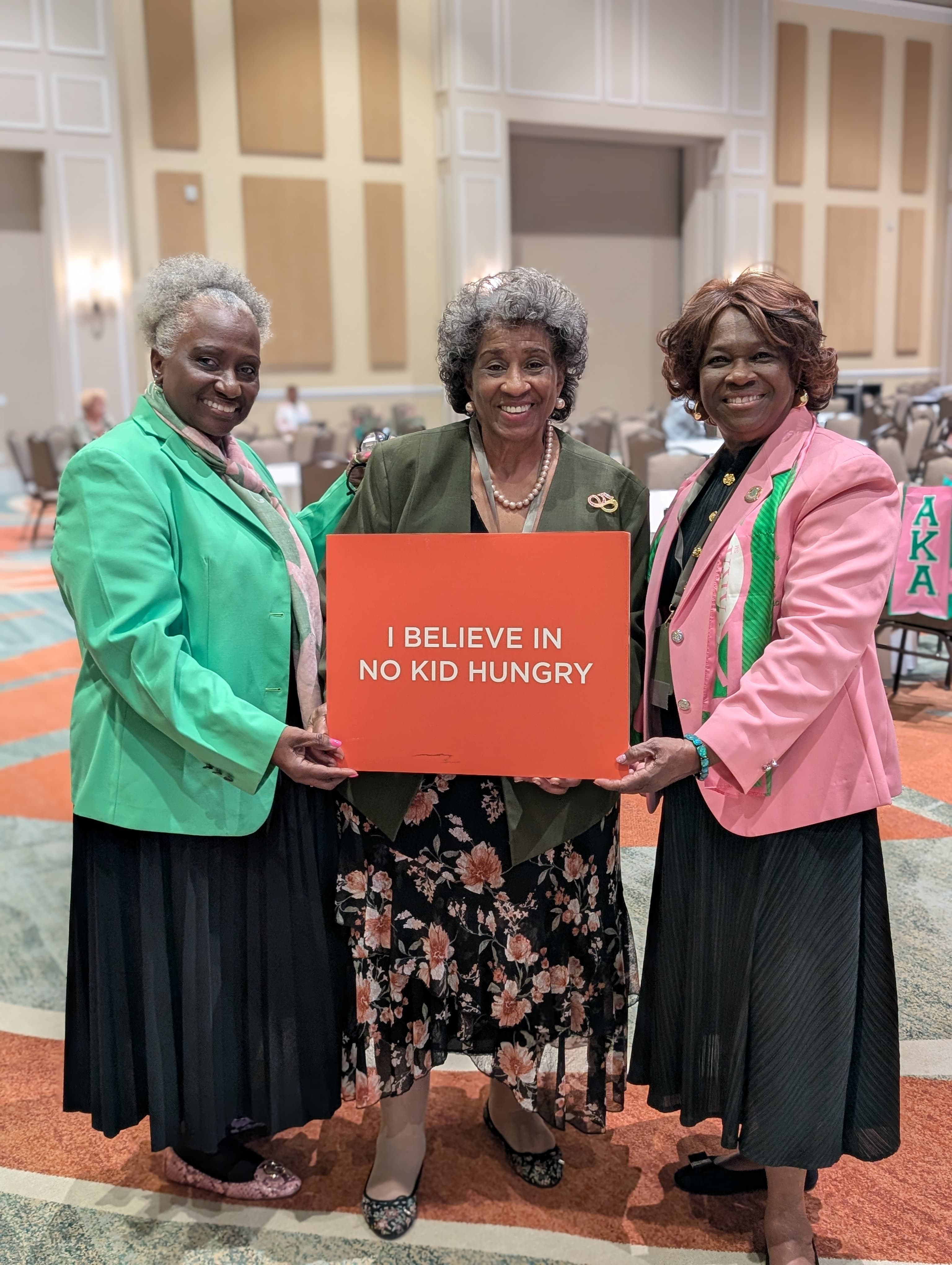 Three AKA members hold a sign that says "I Believe in No Kid Hungry"