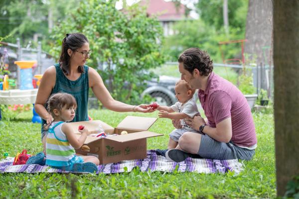 a family of four eats a picnic. The baby sits on their dad's lap while the toddler stares into the distance next to their mom. 