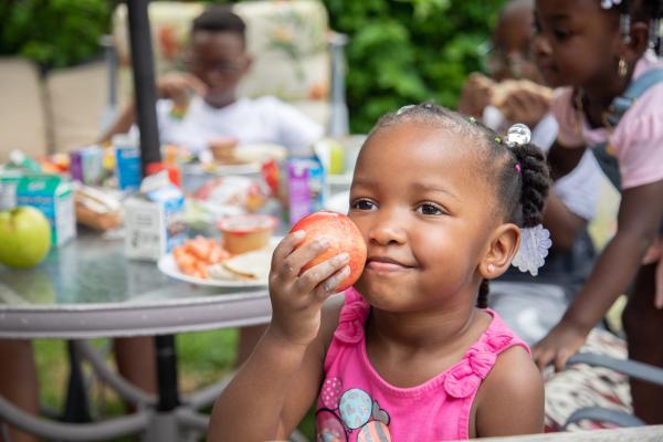 a little girl holds an apple to her face and smiles. In the background two more children are eating their lunch.