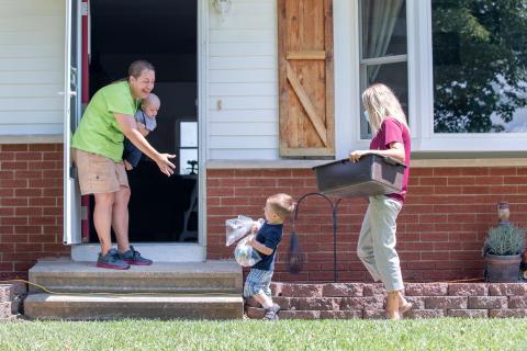 A volunteer brings meals to a family's house and a young boy helps carry them meals inside