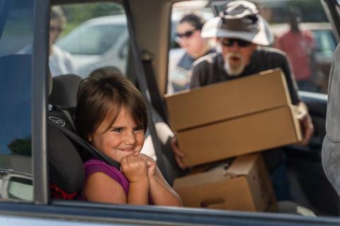 a child smiles out the window while a volunteer loads a box of meals into her family's car
